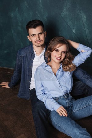 Téléchargez les photos : Jeune homme et femme en jeans vêtements assis ensemble sur un plancher de bois près d'un mur bleu et sourire - en image libre de droit