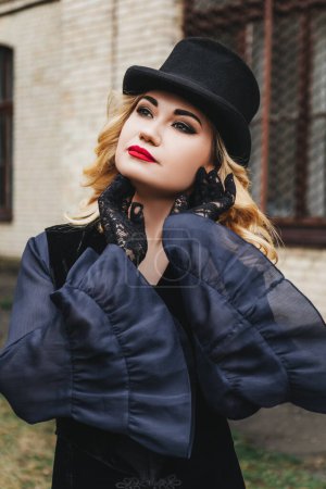 Foto de Hermosa mujer rubia con maquillaje de noche y peinado, labios rojos, en ropa retro, vestido largo azul, guantes guipure y sombrero de copa negro, está de pie cerca de un edificio viejo - Imagen libre de derechos