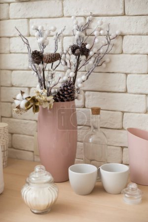 Téléchargez les photos : Un vase rose avec un bouquet de Noël est sur la table de cuisine contre un mur de briques, des branches avec des cônes, du coton et des fleurs d'or, des étincelles, une bouteille transparente avec un bouchon, des tasses, un bol à sucre - en image libre de droit