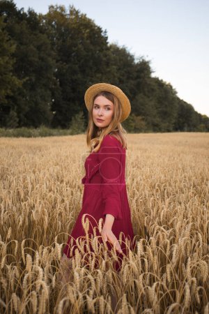 Foto de Mujer joven caminando en el campo de trigo. Señora con sombrero de paja y vestido rojo estilo boho. - Imagen libre de derechos