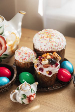 Foto de Platos con tres tradicionales pasteles de Pascua ortodoxos y huevos pintados de rojo, verde y azul, y teteras de flores vintage de pie en la mesa festiva de madera - Imagen libre de derechos