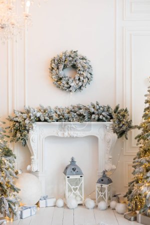 Téléchargez les photos : Intérieur d'une chambre blanche avec cheminée, sapins de Noël avec neige artificielle et guirlandes, canapé, plaid avec oreillers, lanternes, ours en peluche blanc, boîtes avec cadeaux du Nouvel An, une couronne de sapin - en image libre de droit