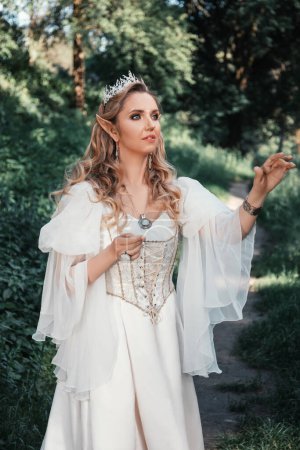 Foto de Hermosa mujer rubia elfa con ojos azules, en un vestido de estilo medieval blanco y en una corona, está de pie en flores blancas, princesa de fantasía - Imagen libre de derechos