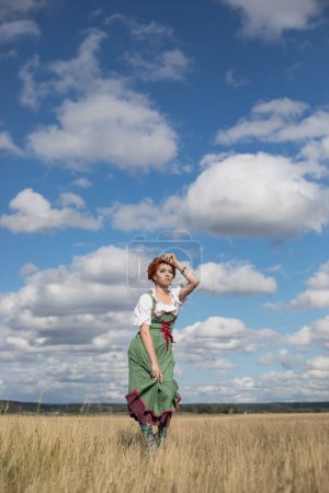Téléchargez les photos : Femme aux cheveux roux en costume national bavarois se tient dans un champ contre un ciel bleu, oktoberfest - en image libre de droit