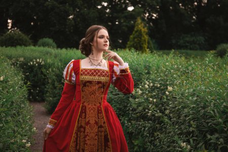 Photo for Beautiful young woman in a red medieval dress is standing in the garden in white flowers, fantasy princess - Royalty Free Image