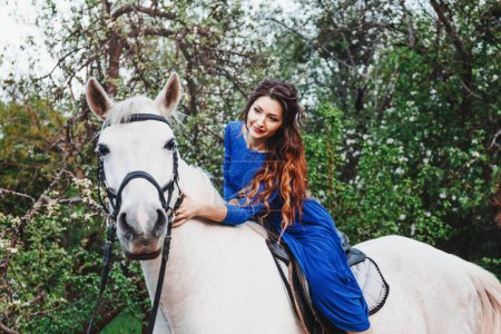Téléchargez les photos : Portrait en plein air printemps d'une belle femme brune sexy aux cheveux bouclés, maquillage du soir, dans une robe bleue et boucles d'oreilles bleues, fleur de pomme, jardin avec arbres, cheval blanc - en image libre de droit