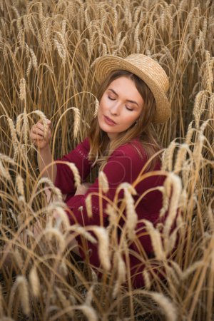 Photo for Young woman sitting in the wheat field and holding a spikelet. Lady wearing straw hat and boho style red dress. - Royalty Free Image