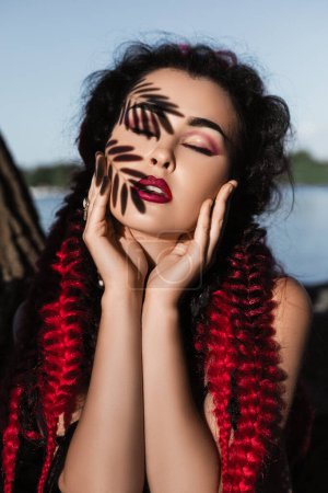Foto de Primer plano retrato de una hermosa joven con el pelo negro, trenzas rojas y maquillaje. sombras de las hojas en la cara femenina - Imagen libre de derechos