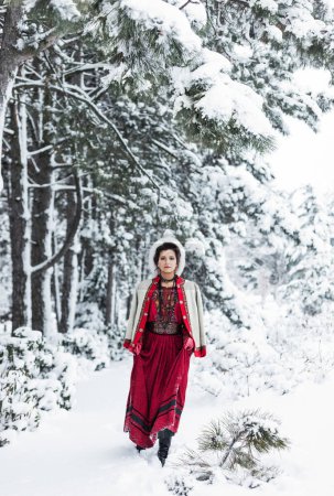 Photo for Attractive brunette woman with evening makeup, in a red ethnic boho style costume with embroidery, white jacket, fur hat, red leather gloves, skirt in a winter snowy forest park - Royalty Free Image