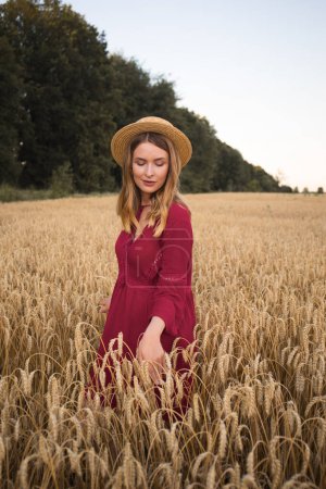 Photo for Young woman standing in the wheat field. Lady wearing straw hat and boho style red dress. - Royalty Free Image