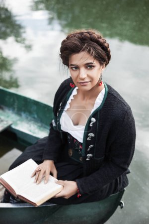 Photo for Portrait of a beautiful brunette woman in a Bavarian folk costume sitting in the boat on the lake and reading a book, braid hairstyle, black skirt, red accessories, countryside, oktoberfest - Royalty Free Image
