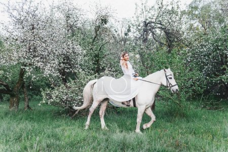 Téléchargez les photos : Portrait en plein air printemps d'une belle femme blonde dans une robe longue blanche et avec des fleurs dans ses cheveux dans un jardin fleuri de pommes chevauchant un cheval blanc, fleurs, tresse, coiffure, maquillage - en image libre de droit