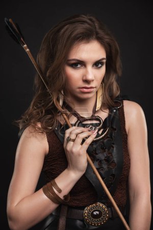 Photo for Beautiful young brunette woman with curly hair and make-up, in boho style clothes and a leather skirt, stands on a black studio background with a bow and an arrow, fantasy warrior, Sagittarius - Royalty Free Image
