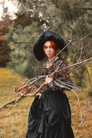 Téléchargez les photos : Femme-sorcière aux cheveux roux en robe noire et chapeau pointu se tient dans la forêt d'automne et tient du broussailles dans ses mains, costume d'Halloween - en image libre de droit
