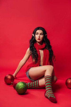 Téléchargez les photos : Jeune femme sexy en costume d'elfe et avec des oreilles elfiques est à l'arrière-plan du studio rouge avec une grosse boule de Noël, style pin-up, nouvelle année vente - en image libre de droit