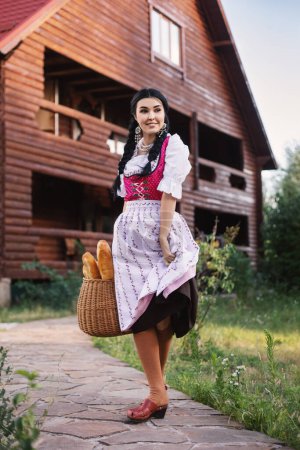 Téléchargez les photos : Belle jeune femme brune en costume bavarois traditionnel se tient près de la maison en bois avec le panier de baguettes, Oktoberfest - en image libre de droit