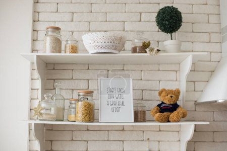 Photo for Brick wall with wooden white kitchen shelves, glass jars with spices and pasta, flower pot, teddy bear - Royalty Free Image