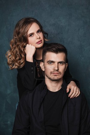 Photo for Young man and woman with makeup and hairstyle in black suits near a blue wall, looking at the camera - Royalty Free Image