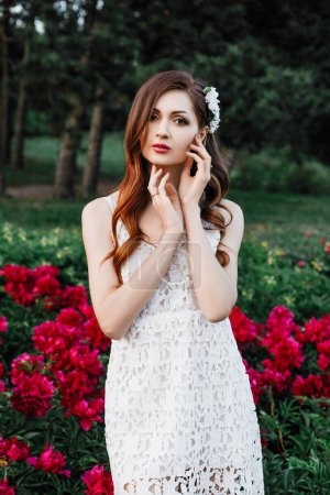 Foto de Retrato de primavera al aire libre de una hermosa mujer morena sexy con el pelo rizado, maquillaje de noche, en un vestido blanco y con flores en el pelo, jardín, flor, peonías rojas - Imagen libre de derechos