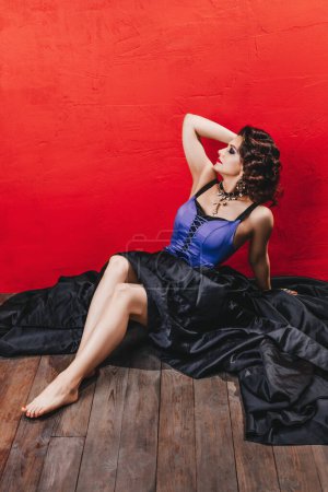 Photo for Beautiful brunette woman with evening makeup and curly hairstyle, red lips, in a black and purple corset dress and with a big cross is sitting on the wooden floor near a red wall, legs - Royalty Free Image
