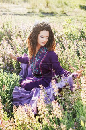 Téléchargez les photos : Jolie femme brune aux cheveux bouclés et au maquillage violet dans une robe ethnique violette et avec couronne de fleurs de lavande est assise en fleur de lavande - en image libre de droit