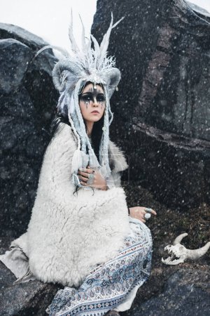 Foto de Hermosa mujer con pelo negro y maquillaje de fantasía, en corona con trenzas, cuernos y plumas, en ropa blanca en estilo étnico boho y en capa de piel sentada en las cañas en la nieve que cae - Imagen libre de derechos