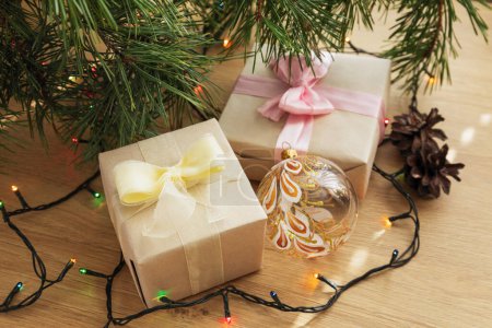 Photo for Christmas gift in craft paper with yellow ribbon, pine branches, cones and a garland on the wooden table - Royalty Free Image