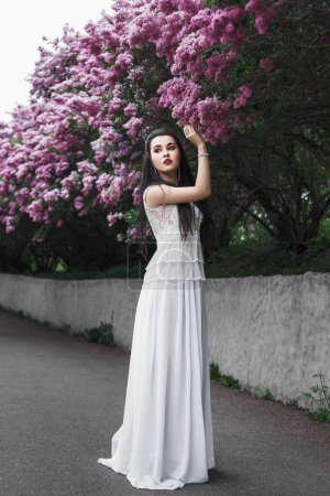 Foto de Retrato de primavera al aire libre de una hermosa mujer morena sexy entre flores lila, vestido blanco largo, pelo negro, maquillaje de noche, joyas, jardín, flor - Imagen libre de derechos