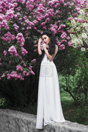 Foto de Retrato de primavera al aire libre de una hermosa mujer morena sexy entre flores lila, vestido blanco largo, pelo negro, maquillaje de noche, joyas, jardín, flor - Imagen libre de derechos
