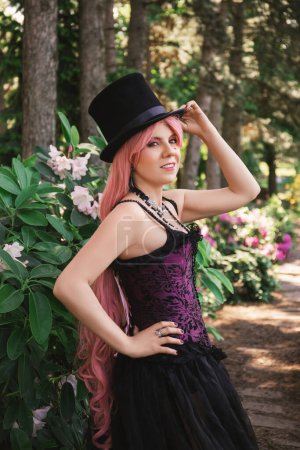 Foto de Hermosa mujer con el pelo rosa, en un sombrero de copa y en un vestido negro de noche con un corsé en el jardín en flores de azalea - Imagen libre de derechos