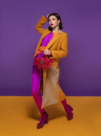 Téléchargez les photos : Belle femme brune avec maquillage de couleur dans une robe violette et une ceinture orange, dans un manteau jaune et des chaussures rouges, avec un sac rouge et une boucle d'oreille de plumes jaunes se tient sur un studio violet et jaune - en image libre de droit