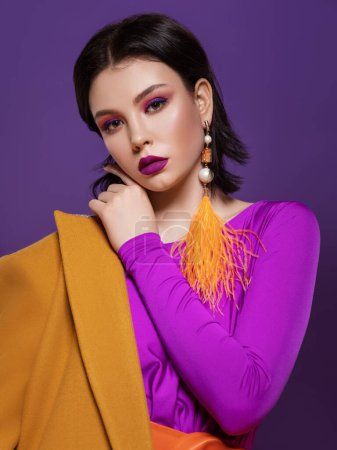 Photo for Beautiful brunette woman with color make-up in a purple dress and an orange belt, in a yellow coat and red shoes, with a red bag and an earring of yellow feathers stands on a purple and yellow studio - Royalty Free Image