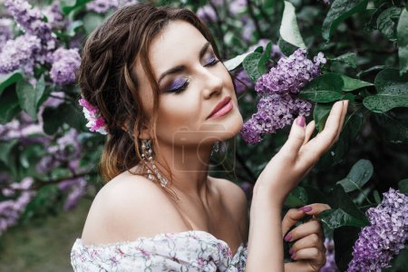 Photo for Spring portrait of a beautiful brunette woman with curly hair, in a long light dress and in a crown, sitting in flowers among the branches of lilac bloom - Royalty Free Image