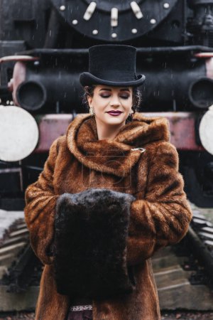 Téléchargez les photos : Portrait de style rétro de belle femme brune en costume vintage et chapeau près du vieux train, manteau de fourrure, tresses de couleur, maquillage du soir, yeux bleus, pluie - en image libre de droit
