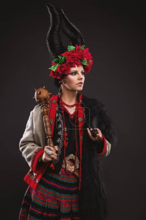 Photo for Beautiful brunette woman in a traditional Ukrainian costume, with a floral wreath and horns, is standing on the black studio background - Royalty Free Image