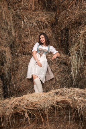 Photo for Beautiful young brunette woman in a traditional bavarian costume is sitting on the hayloft and smiling, oktoberfest - Royalty Free Image