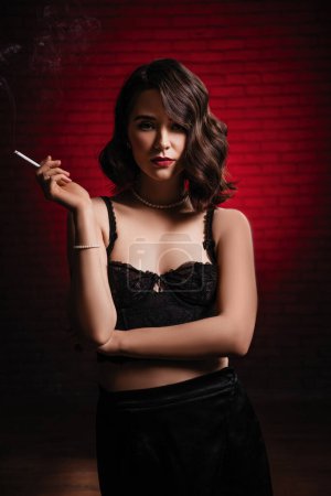 Foto de Sexy joven morena con maquillaje de noche y pelo rizado, labios rojos, en un vestido de corsé guipur negro y medias de pie sobre el fondo de la pared de ladrillo rojo, negro, tocador, primer plano - Imagen libre de derechos
