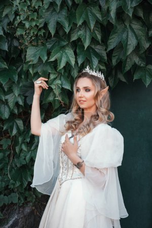Téléchargez les photos : Belle femme elfe blonde aux yeux bleus, dans une robe de style médiéval blanc et dans une couronne, est debout dans des fleurs blanches, princesse fantaisie - en image libre de droit