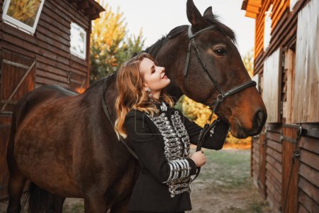 Téléchargez les photos : Belle femme modèle en costume noir est debout avec un cheval près de l'écurie - en image libre de droit