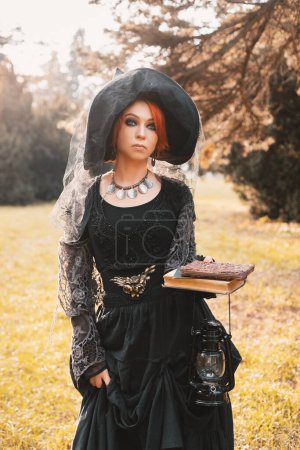 Foto de Una bruja pelirroja en el vestido negro y sombrero apuntado en un parque de otoño y sonriente, disfraz de Halloween - Imagen libre de derechos