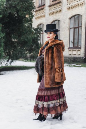 Téléchargez les photos : Portrait de style rétro de belle femme brune en costume vintage et chapeau près du vieux bâtiment, manteau de fourrure, tresses de couleur, maquillage du soir, yeux bleus, neige - en image libre de droit