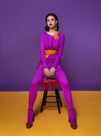 Téléchargez les photos : Belle femme brune avec un maquillage de couleur vive dans une robe violette et une ceinture orange, en rouge à carreaux chaussures à talons hauts, avec un sac rouge et une boucle d'oreille en plume jaune assis sur une chaise - en image libre de droit