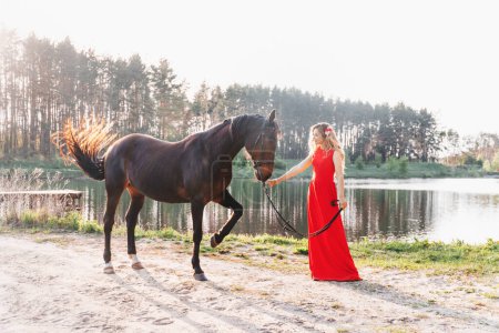 Téléchargez les photos : Printemps été portrait en plein air d'une belle femme blonde dans une robe longue rouge et avec des fleurs dans ses cheveux debout dans la forêt au bord du lac avec un cheval, coiffure, maquillage - en image libre de droit
