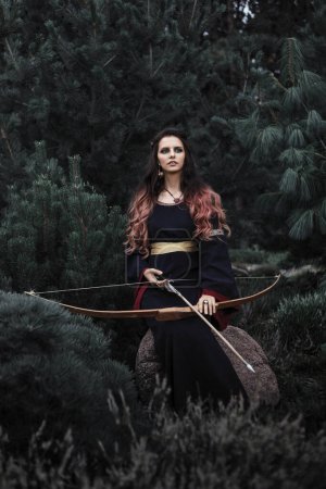 Foto de Hermosa princesa elfa oscura en el bosque. Bruja de fantasía con arco y flecha. - Imagen libre de derechos