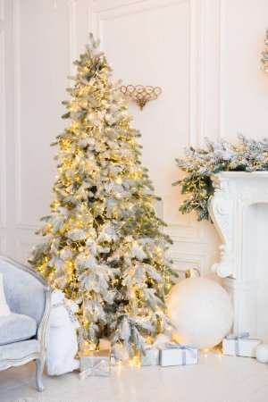 Téléchargez les photos : Intérieur d'une chambre blanche avec cheminée, sapins de Noël avec neige artificielle et guirlandes, canapé, plaid avec oreillers, lanternes, ours en peluche blanc, boîtes avec cadeaux du Nouvel An, une couronne de sapin - en image libre de droit