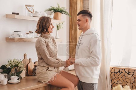 Photo for Cheerful young couple talking in the kitchen. Happy woman sitting on the table. Husband and wife holding hands. Couple spending time together at home. - Royalty Free Image