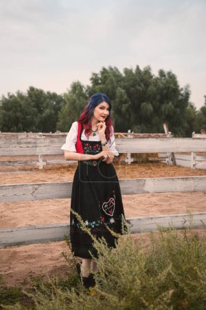 Photo for Beautiful young woman in a traditional bavarian costume is at the fence on the farm and smiling, oktoberfest - Royalty Free Image