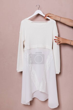 Photo for Female hands with manicure and bracelets are holding a hanger against a pink wall with a white dress with chiffon and lace - Royalty Free Image