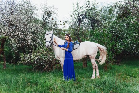 Foto de Retrato de primavera al aire libre de una hermosa mujer morena sexy con el pelo rizado, maquillaje de noche, en un vestido azul y flores blancas en su cabello, flor de manzana, jardín con árboles, caballo blanco - Imagen libre de derechos