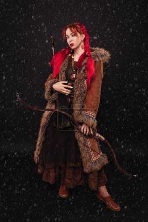 Photo for Pretty woman with blue eyes and red hair, in a brown fur coat and dress in ethnic style, with a bow in her hands stands on the black background in the falling snow - Royalty Free Image